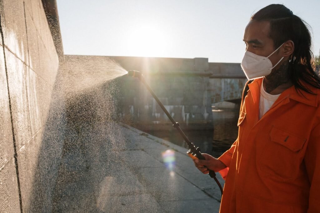 Man in orange uniform wearing facemask sprays water on wall outdoors, sunlight shining.