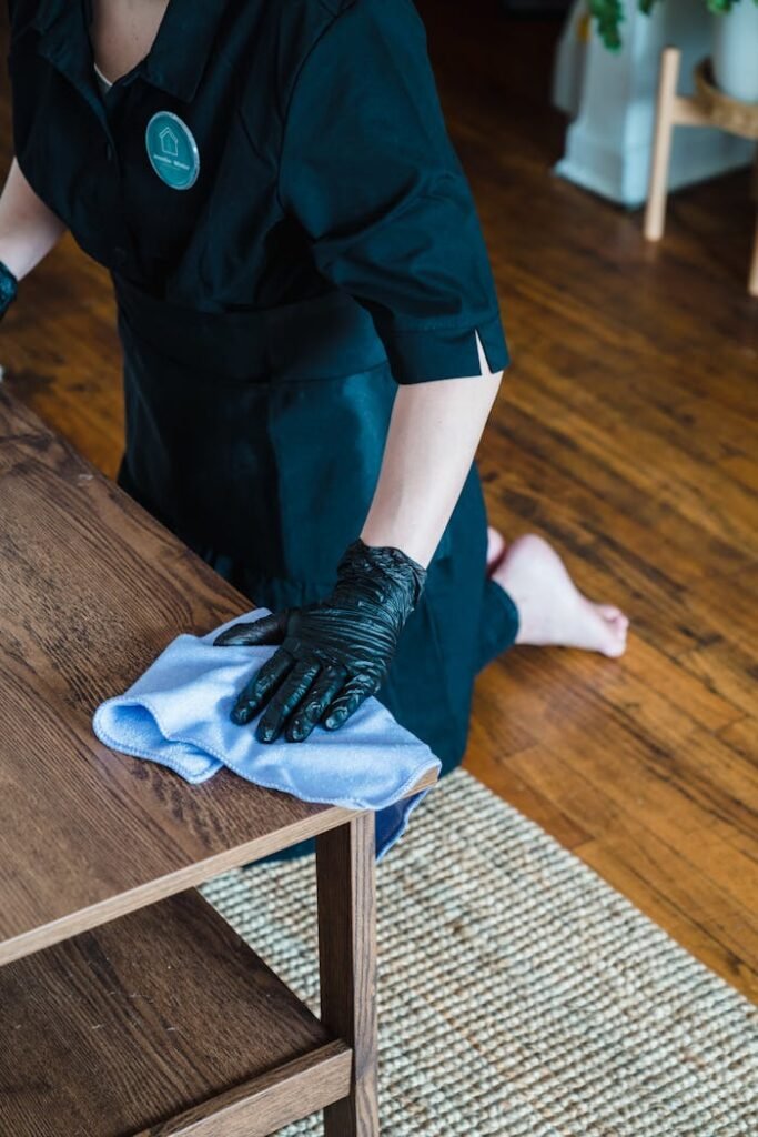 A professional cleaner wearing gloves and uniform wipes a wooden table surface indoors.
